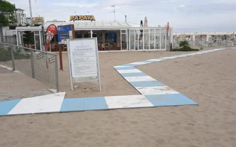 Percorso a strisce bianche e blu sulla sabbia del piazzale San Martino a Riccione, conduce al bar Papaya e alla spiaggia attrezzata.