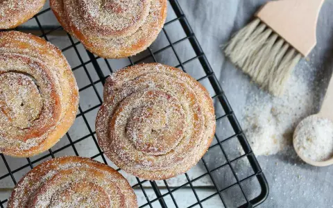 Nybakade karlsbaderbullar med socker och kanel på ett svart galler. En borste och en sked med socker ligger bredvid.
