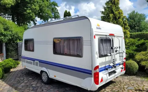 Biała przyczepa kempingowa Hobby 440 z niebieskimi pasami, zaparkowana na brukowanym podjeździe. Idealna do podróżniczego hobby.