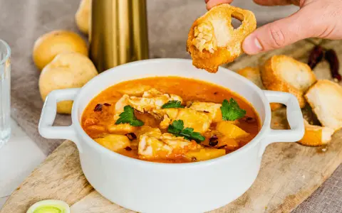 Una mano intinge un crostino nella zuppa di pesce surgelato, ricca di sapori e profumi.