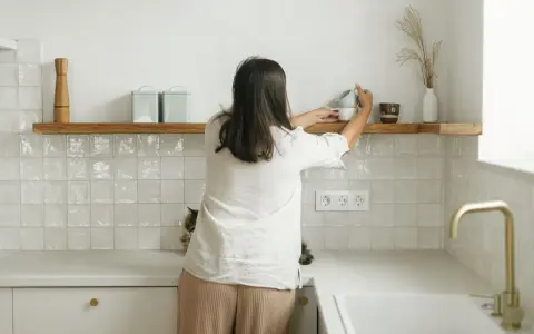 Kobieta układa kubki na drewnianych, otwartych półkach w kuchni. Obok kotek obserwuje porządki.