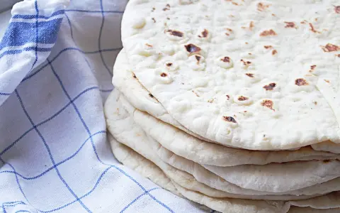 En hög med nybakade tortillabröd, perfekta för ditt nästa tortilla recept. Bredvid ligger en blåvitrutig kökshandduk.