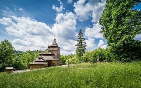 Drewniana cerkiew na tle błękitnego nieba z chmurami, otoczona zielenią. Widok z łąki w Beskidzie Dolnym.