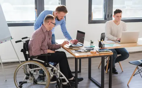 Współpraca w biurze: mężczyzna na wózku inwalidzkim, z umiarkowanym stopniem niepełnosprawności, pracuje z kolegą przy laptopie.