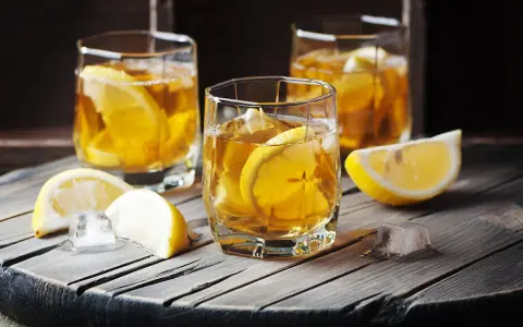 Metaxa orange z czym pić? Idealnie z lodem i plasterkiem cytryny. Trzy szklanki złocistego trunku na drewnianym blacie.