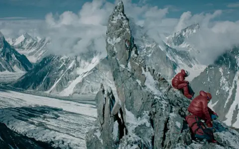 Dwóch alpinistów w czerwonych kurtkach odpoczywa na skalistej grani, podziwiając rozległy widok na lodowiec i ośnieżone szczyty. Broad peak góra w oddali.