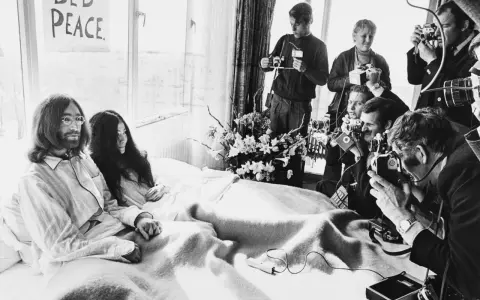 John Lennon i Yoko Ono w łóżku, otoczeni przez reporterów z aparatami. Na ścianie wisi plakat "BED PEACE".