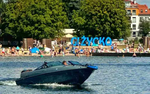 Motorówka sunie po wodzie, w tle plaża pełna ludzi i wielki napis "GIŻYCKO". Idealny dzień na Mazurach.