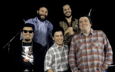 Los Lobos, native sons, pose together. One man wears sunglasses and a fringed jacket, another a denim jacket, and two sport plaid shirts. A saxophone is visible.
