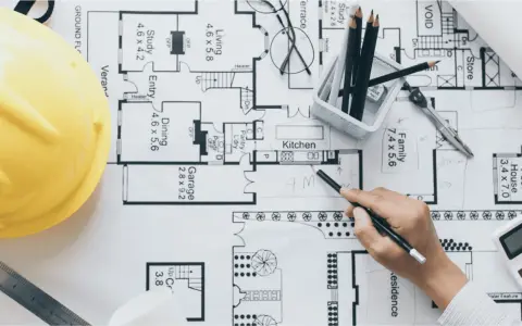 Un arquitecto o proyectista de una obra revisa planos detallados de una casa, con casco amarillo, lápices y calculadora.