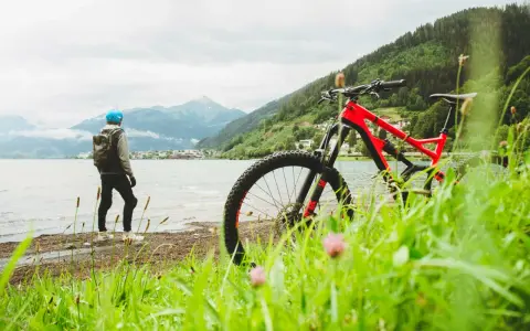 En person med hjälm och ryggsäck står vid en sjö med en mountainbike framför sig. Berg och en stad syns i bakgrunden. Perfekt för att cykla MTB.
