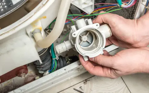 Hände halten eine Waschmaschine-Pumpe, die nicht abpumpt. Kabel und Schläuche sind sichtbar.