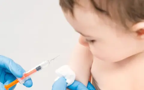 Dłoń w niebieskiej rękawiczce trzyma strzykawkę, przygotowując się do szczepienia niemowlęcia.