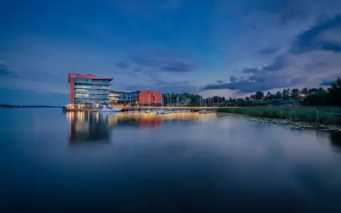 Nowoczesny hotel na ptasiej wyspie, otoczony spokojną wodą i zielenią, z łodziami przy pomoście.