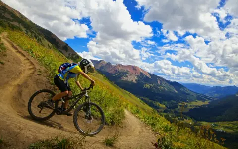 A mountain biker navigates a steep, winding trail, experiencing the best mountain biking in Colorado with stunning valley views and dramatic clouds.