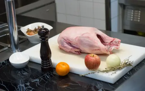 Rohe Gans auf einem Schneidebrett mit Apfel, Zwiebel, Orange, Gewürzen und Pfeffermühle. Die Frage "wie lange dauert auftauen" beschäftigt jeden Koch.