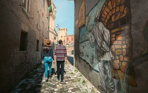 Coppia passeggia in un vicolo di Saludecio, ammirando un murale. Scopri cosa vedere in questo borgo incantevole.