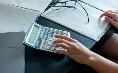 Ręka wpisuje dane na kalkulatorze, przygotowując się do zajęcia wierzytelności. Obok leżą dokumenty i okulary.