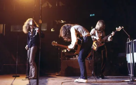 Energetyczny **Led Zeppelin koncert** na scenie. Wokalista z burzą włosów, gitarzysta pochylony nad instrumentem, basista grający w tle.