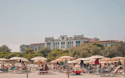 Entspannte Menschen liegen unter Sonnenschirmen am Lido di Venezia Beach. Im Hintergrund thront ein imposantes Gebäude vor üppigem Grün.