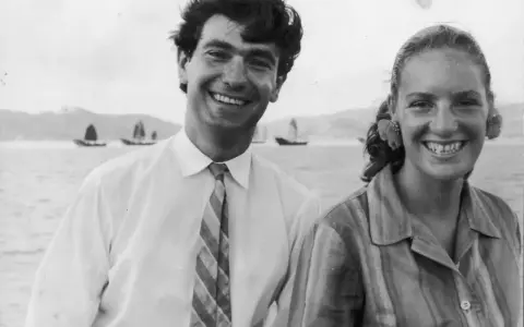 Angela Staude sorride con un uomo su una barca, con velieri sullo sfondo.