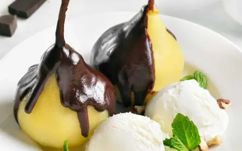 Poires pochées nappées de chocolat noir, servies avec de la glace à la vanille et des éclats de noisettes. Un délice de poire Belle Hélène.