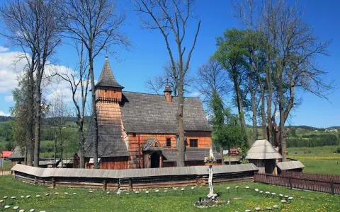 Drewniany kościół w Dębnie, otoczony kamiennym murem i drzewami, z krzyżem misyjnym na trawiastym dziedzińcu.