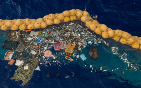 Ogromna wyspa śmieci na oceanie, zebrana w siatkę z bojami. Plastikowe odpady, sieci rybackie i opona tworzą przygnębiający widok.
