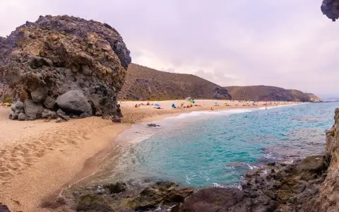 Plaża w Cabo de Gata z turkusową wodą, piaszczystym brzegiem i skalistymi formacjami. Ludzie odpoczywają pod parasolami.