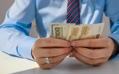 Mężczyzna w niebieskiej koszuli i krawacie liczy banknoty 100 i 200 zł. Może to być związane z rozliczeniem zus preferencyjny.