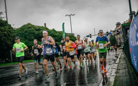 Uczestnicy maratonu biegną w deszczu po mokrej nawierzchni. Widać ich skupienie i determinację mimo niesprzyjającej pogody.