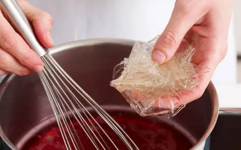 Hände bereiten Gelatine-Blätter für die Zubereitung vor, indem sie sie in eine rote Flüssigkeit in einem Topf geben, während ein Schneebesen bereitsteht.