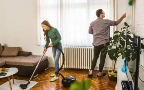 Pareja colaborando para purificar casa. Ella aspira el suelo, él quita el polvo de las paredes.