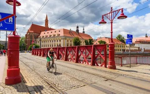 Mężczyzna na rowerze jedzie po brukowanej drodze rowerowej z torami tramwajowymi, w tle zabytkowe budynki i czerwony most.