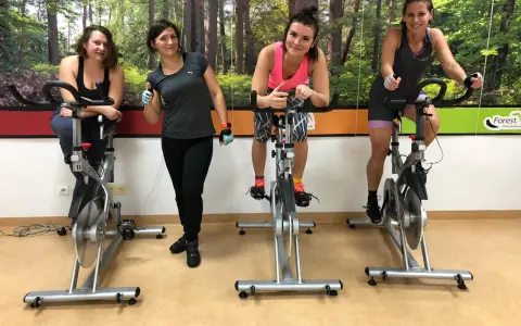 Cztery kobiety ćwiczą na rowerach stacjonarnych. W tle las. To świetny sposób na trening spinning co to jest?