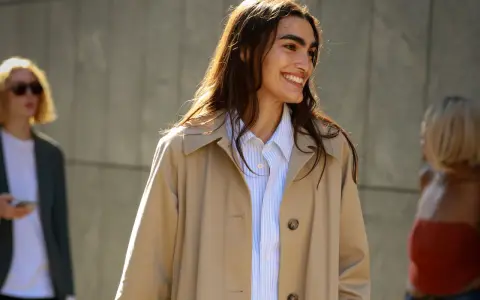 Donna sorridente con capelli lunghi e castani indossa un trench beige aperto su una camicia a righe azzurre. Perfetto per ogni occasione, anche se posso mettere il trench di sera.