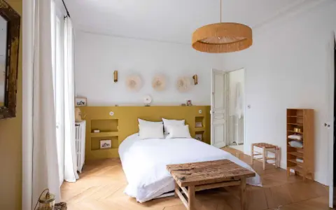Une chambre lumineuse avec une tête de lit peinture jaune moutarde, des décorations murales en paille et un lit blanc douillet.