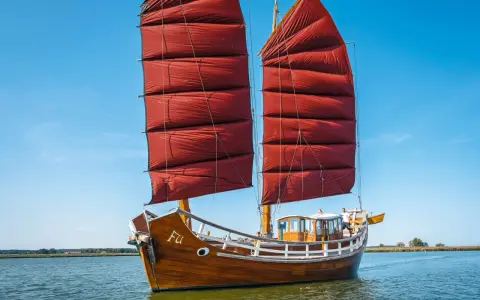 Ein hölzernes Dschunke Schiff mit roten Segeln gleitet auf ruhigem Wasser unter blauem Himmel.