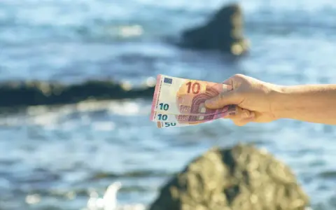 Dłoń trzyma banknoty euro na tle morza. W Czarnogórze obowiązuje euro, więc te pieniądze przydadzą się na wakacjach.