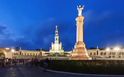Pilgrims gather at the Sanctuary of Fátima, Portugal, at dusk, with the illuminated basilica and statue of Christ the King standing majestically.
