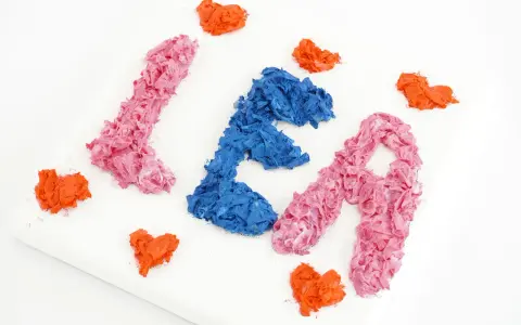 Comment faire du relief sur un tableau : des lettres "LÉA" en papier froissé rose et bleu, entourées de cœurs orange sur une toile blanche.