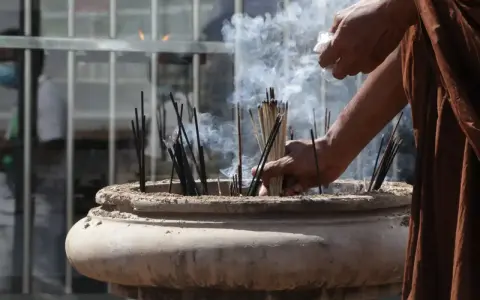 Manos monje en túnica marrón colocando varillas de incienso en un cuenco de piedra, creando humo para limpiar el ambiente.