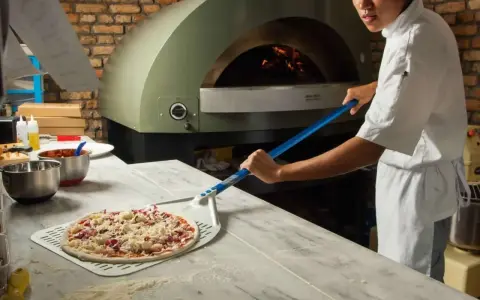 Pizza cruda en pala, lista para horno de leña con chef al fondo.