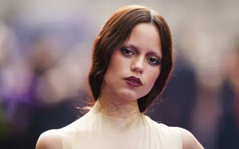 A young woman with auburn hair and a dramatic, dark lipstick looks directly at the camera. Her eye makeup is smudged, giving her a tired girl makeup look, as if she's been out all night.