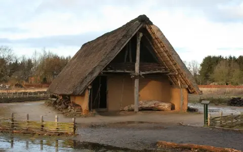 Ein rustikales Bauernhaus aus dem Mittelalter mit Strohdach und Lehmwänden, umgeben von Holzzäunen und Bäumen.