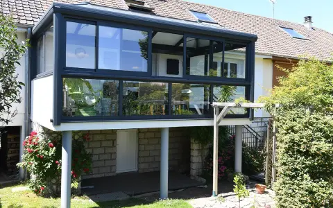Moderne veranda adossée au mur, avec une chaise verte visible à travers les grandes baies vitrées. Le soleil brille sur le jardin verdoyant.