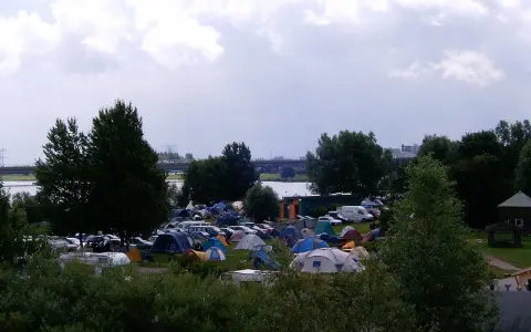 Rozległy kamping z mnóstwem namiotów i samochodów, nad wodą, pod pochmurnym niebem.