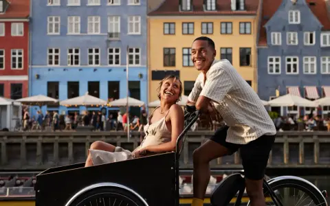 Ett par njuter av en romantisk weekend i Europa, cyklar längs en kanal med färgglada hus i bakgrunden.
