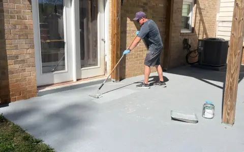Un homme applique une peinture sur mur béton extérieur avec un rouleau. Le sol gris est en cours de rénovation.