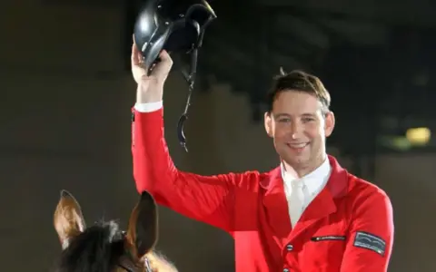 Reiter Franke Sloothaak hält stolz seinen Helm hoch, trägt eine rote Reitjacke und sitzt auf einem braunen Pferd.